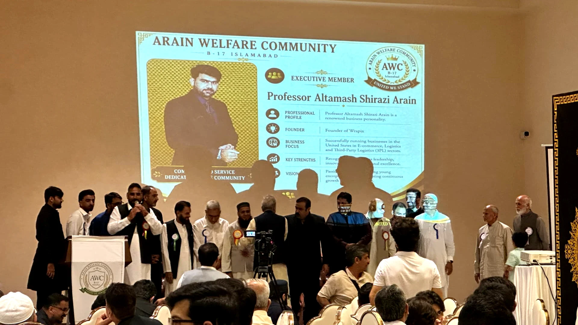 Executive member profile displayed on the projection screen while members stand on stage during the Arain Welfare Community annual gathering